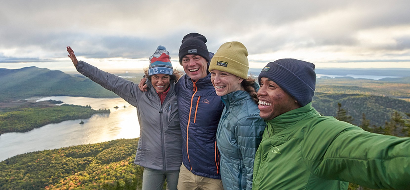 4 smiliing friends, arms around each other and outstretched, on a beautiful mountain top.