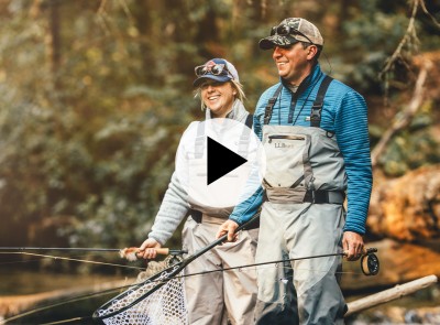 A smiling man and woman standing knee deep in water with fly fishing equipment.