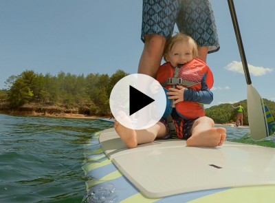 Toddler in a life jacket sitting on a stand up paddle board with a parent.