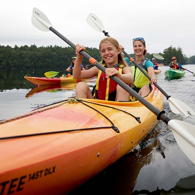 Two people smiling as they paddle in kayaks towards the screen