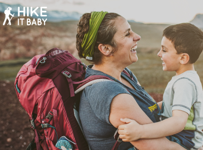 Laughing mom with backpack holding her happy child on a hike.