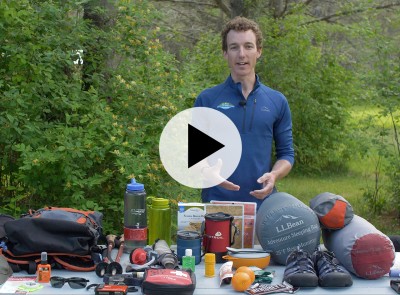 An L.L.Bean instructor behind a table covered with things to consider bringing on a hike, a play video icon in the center.