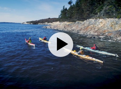 4 kayakers paddling on the ocean near the rocky shore, a play video icon in the center.