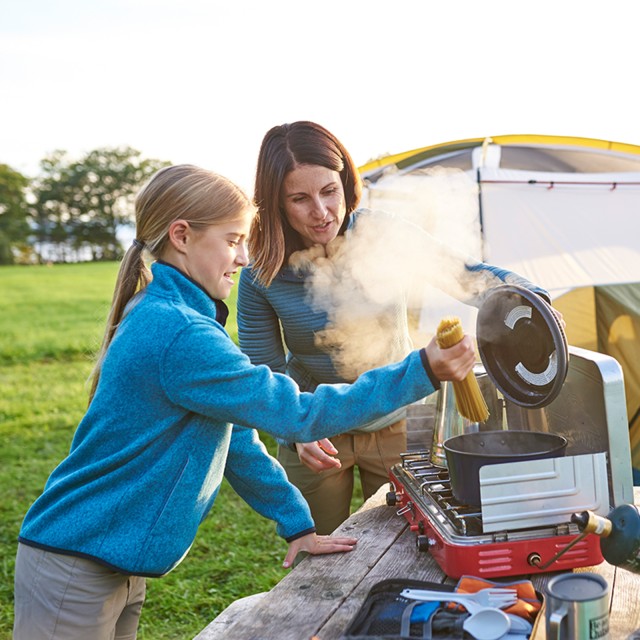 A woman and young girl put pasta into a pot on a portable camp stove.