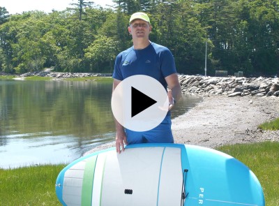 A man holding a SUP by the ocean, a play video icon in the center.