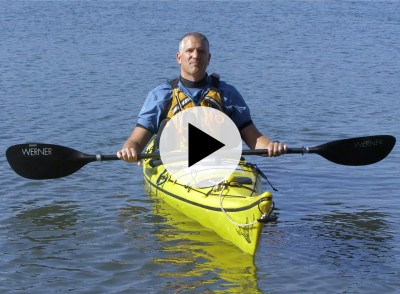 A man holding a kayak paddle sitting in a kayak on the water, a play video icon in the center.