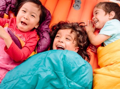 Laughing children cuddled inside sleeping bags in a tent.
