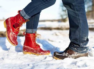 Two sets of Bean Boot clad feet facing each other in the snow, one red Bean Boot raised as if they are kissing.