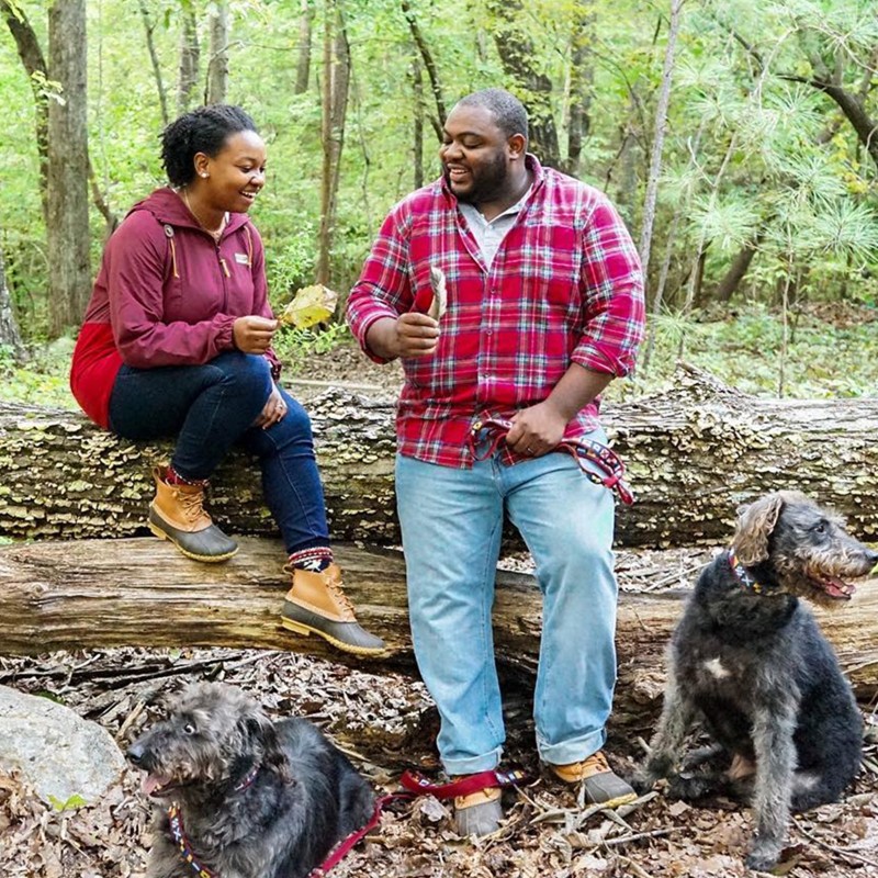 A couple resting on a log in the woods, their 2 dogs sitting at their feet.