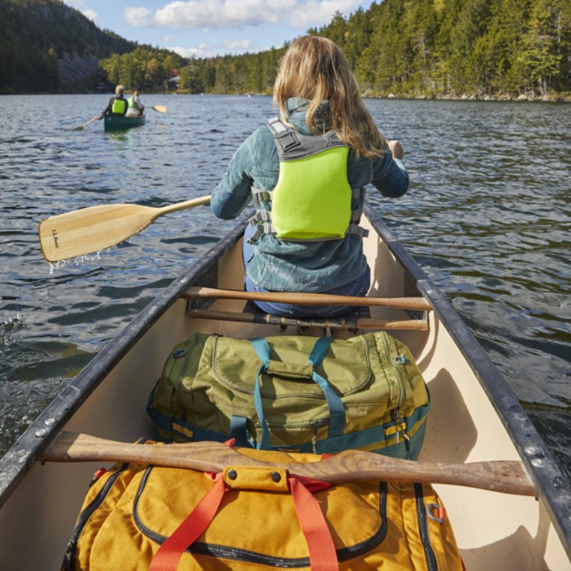 A woman paddling a canoe, shot from behind her, and another canoe with 2 paddlers ahead in the distance.