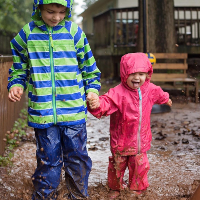 2 kids in rain jackets and pants walking through a mud puddle in the rain.