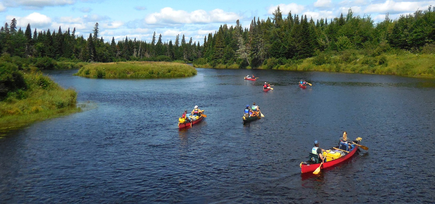 A group of 6 canoes loaded with camping gear and 12 people paddling on calm water on a beautiful day.