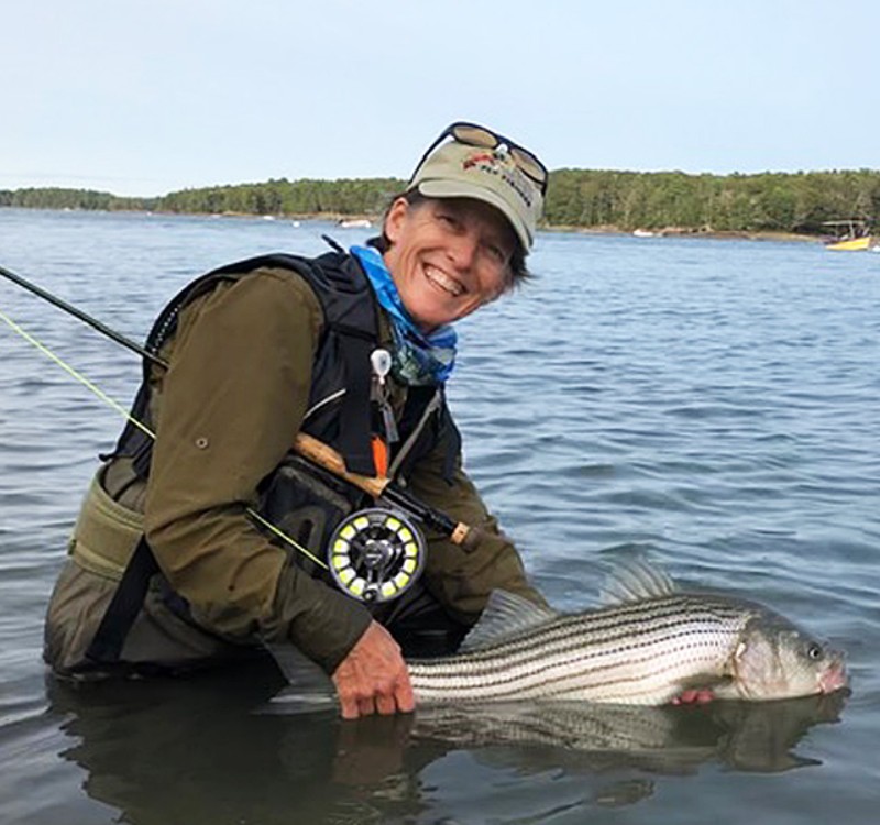 Susan Daignault, with fishing rod under her arm, smiles while standing in thigh-deep water holding a fish.