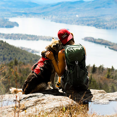 A woman with her arm around a dog looking at a magnificent mountain view.