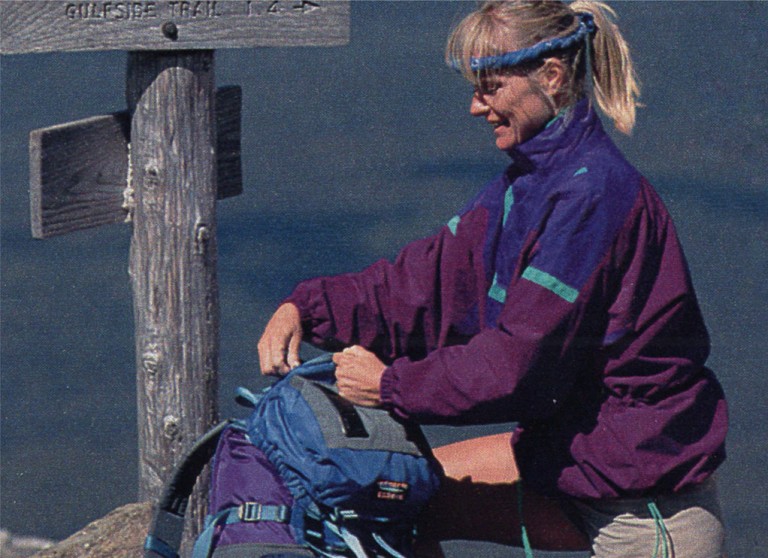 Woman in anorak adjusting her backpack by a trail sign.