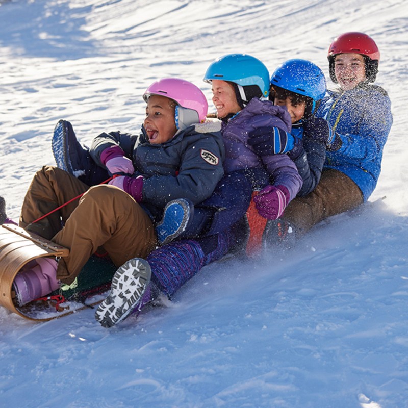 4 happy kids in helmets tobagganing down a snowy hill.