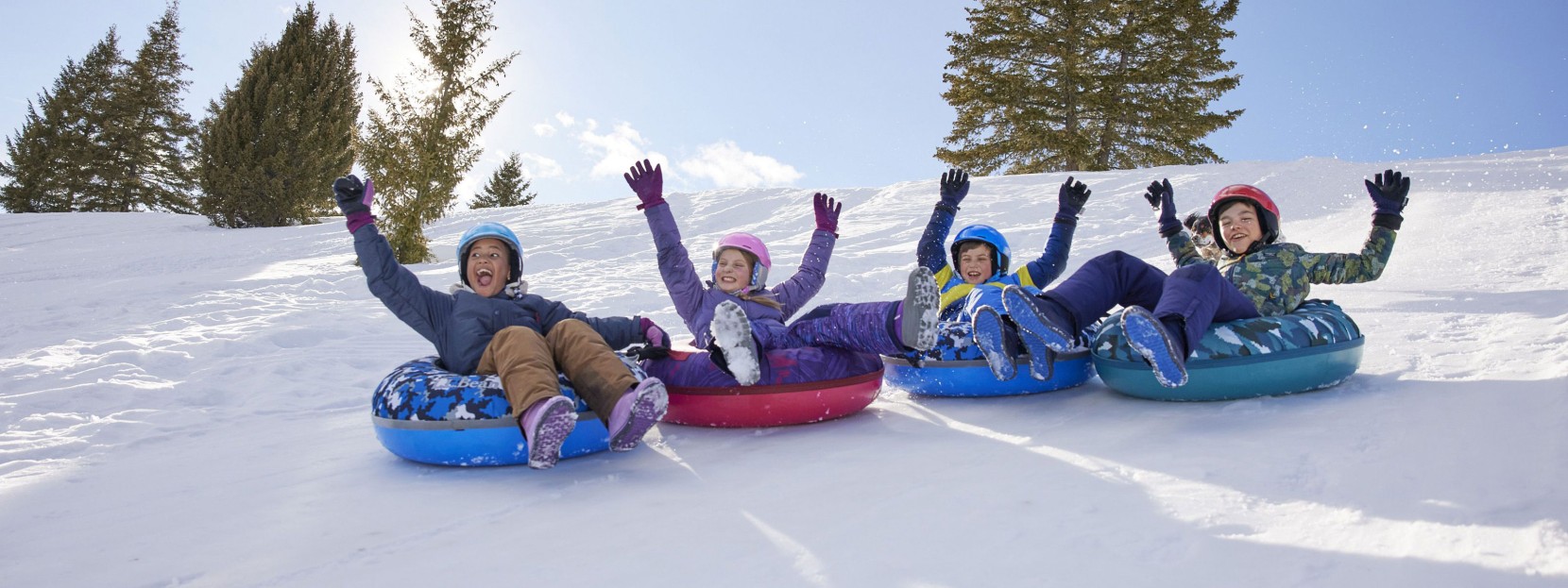 4 kids snow tubing together down a hill having fun with their hands in the air.