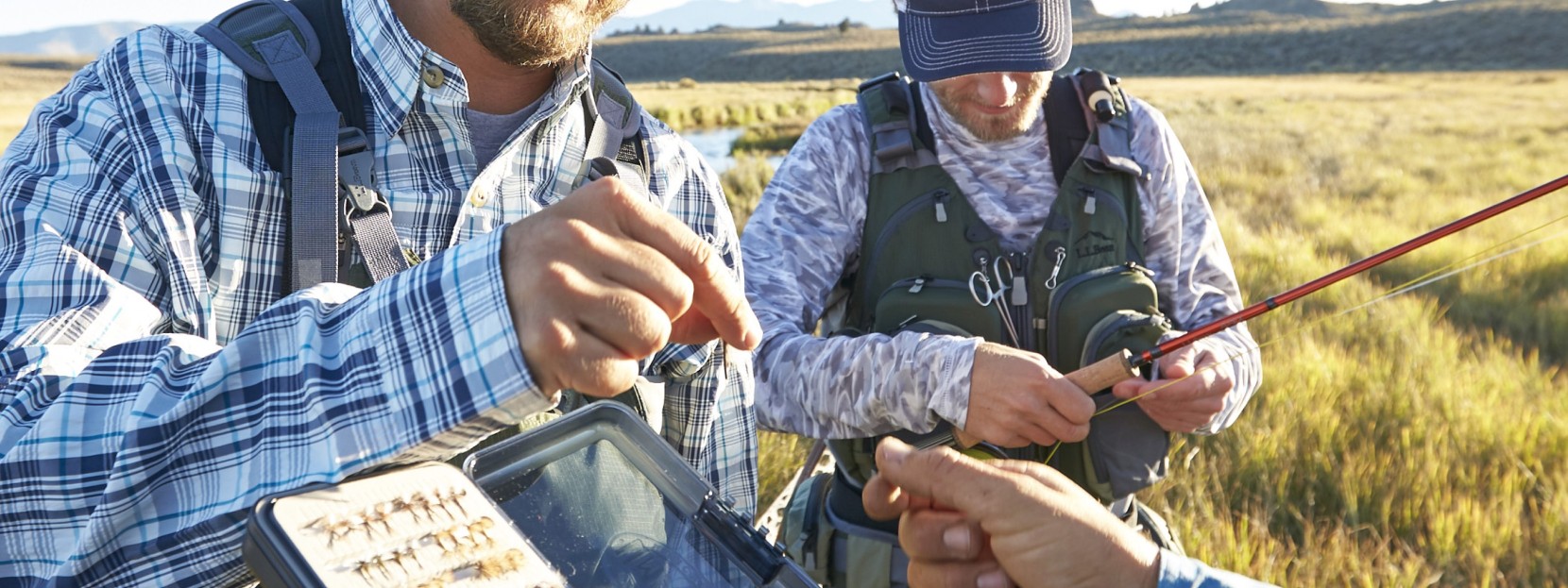 One man holds a fishing rod while two others select a fly from a box with an assortment of them inside