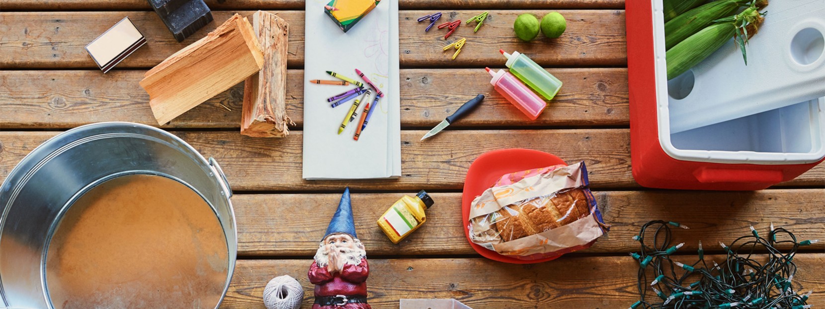 An outdoor table strewn with things to help create a fun backyard party, from a cooler with corn on the cob to string lights.