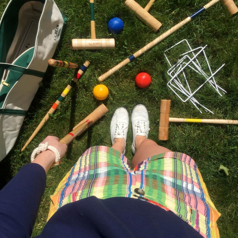 Looking down on someone's legs and feet with parts of a croquet set scattered on the grass.