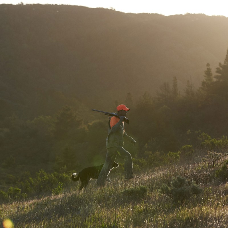 A man with a rifle on his shoulder and a dog walking through a field.