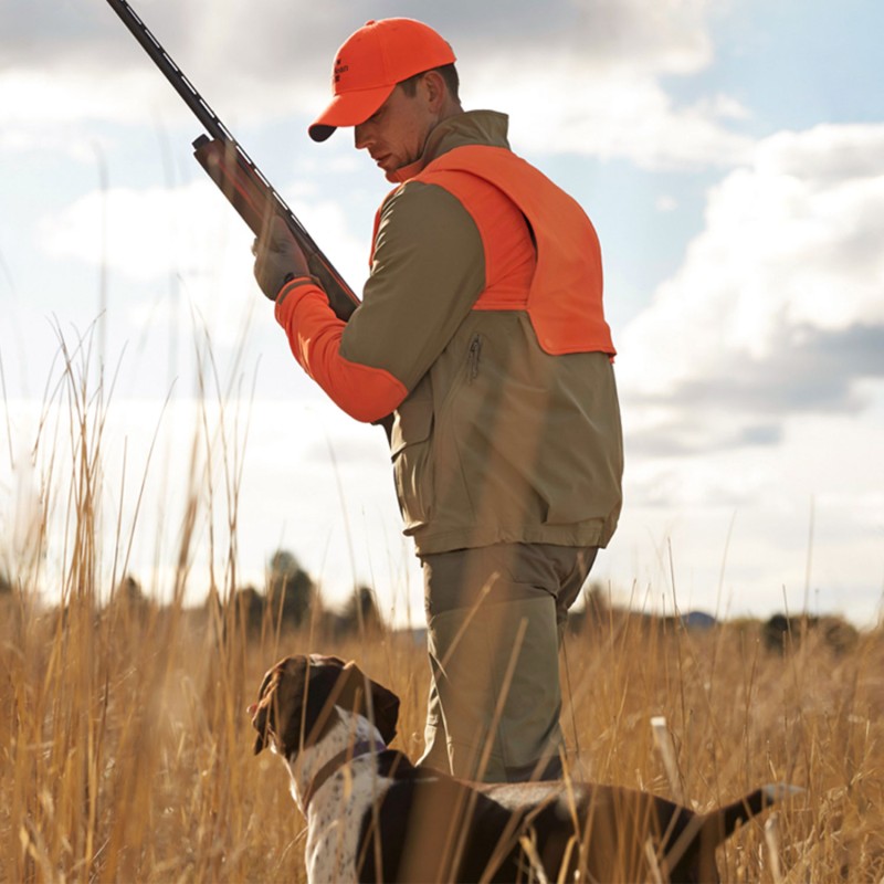 A man with a rifle and wearing a hunter's orange jacket and hat, looks at his dog by his side.