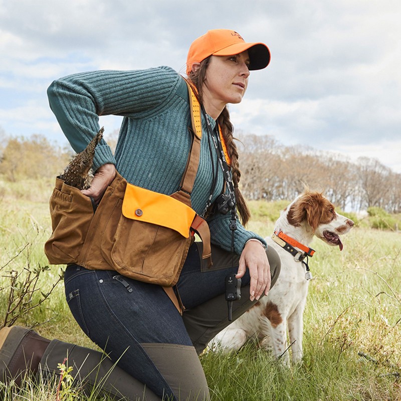 A woman hunter kneeling the field next to a dog, both looking intently into the distance.