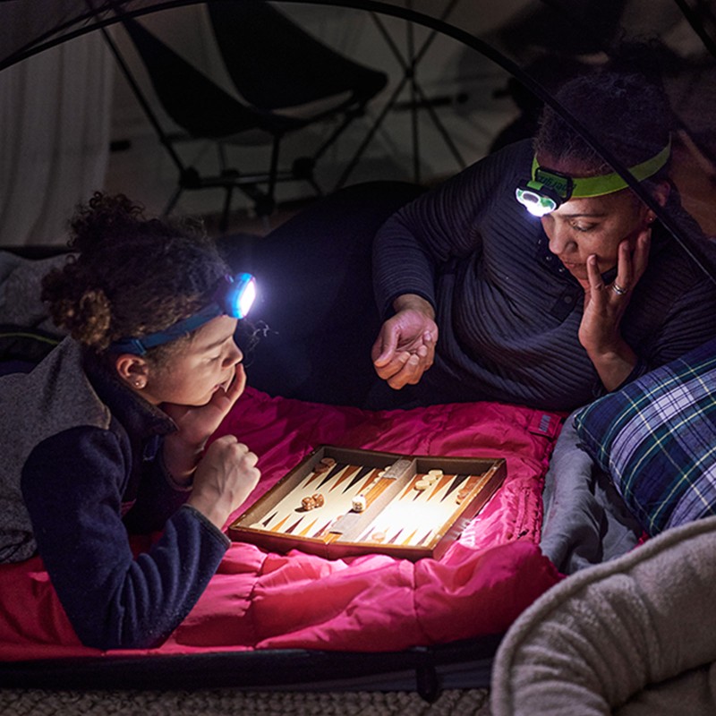 Mom and child with headlamps on, playing backgammon inside a tent.