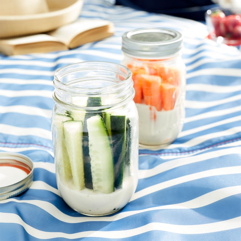 Close-up of a picnic blanket on the grass, 2 mason jars containing cucumbers, carrots & dip, and other typical picnic items.