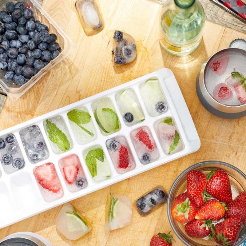 Ice cubes containing blueberries, mint leaves, strawberries or lime slices in an ice cube tray on a kitchen counter.