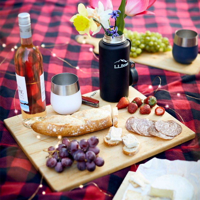 Beautiful outdoor picnic spread on blanket, including a cutting board with baguette, cheese, grapes and wine.