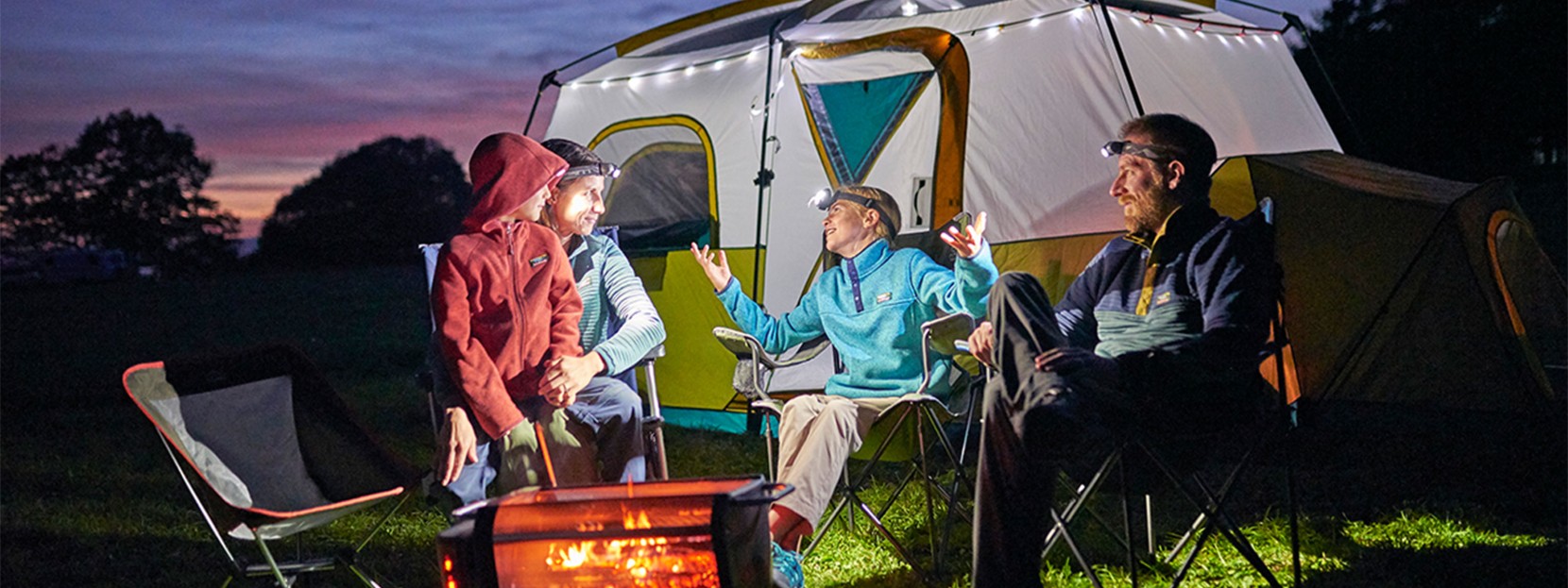 Happy family wearing headlamps sitting around a camp fire with their tent in the background.