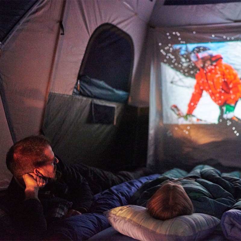 A family lounging in sleeping bags, watching a movie projected on a tent wall.