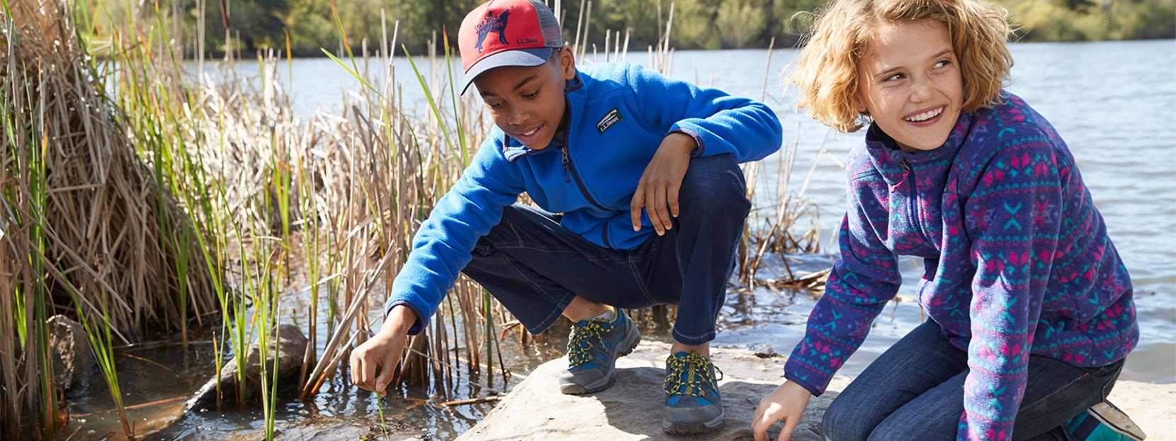 A boy and a girl kneel on a rock in a lake, looking in the water.