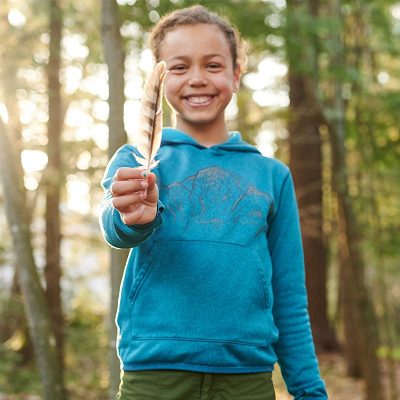 A smiling girl outside in the woods, holding a feather up and looking at the camera.