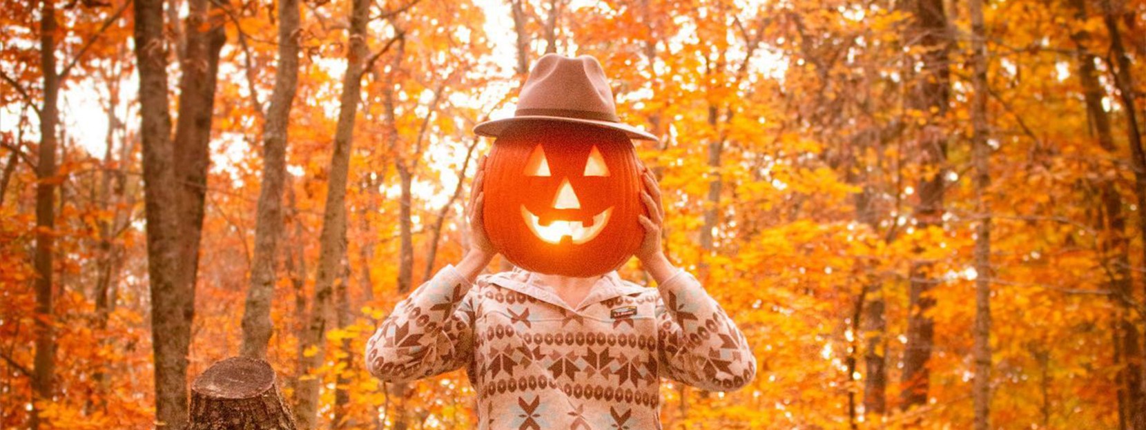 Woman outside holding a lit jack-o'lantern with hat in front of her head.