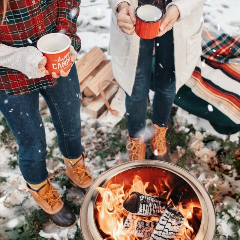 Looking down on 2 people enjoying a hot beverage around a firepit in winter.