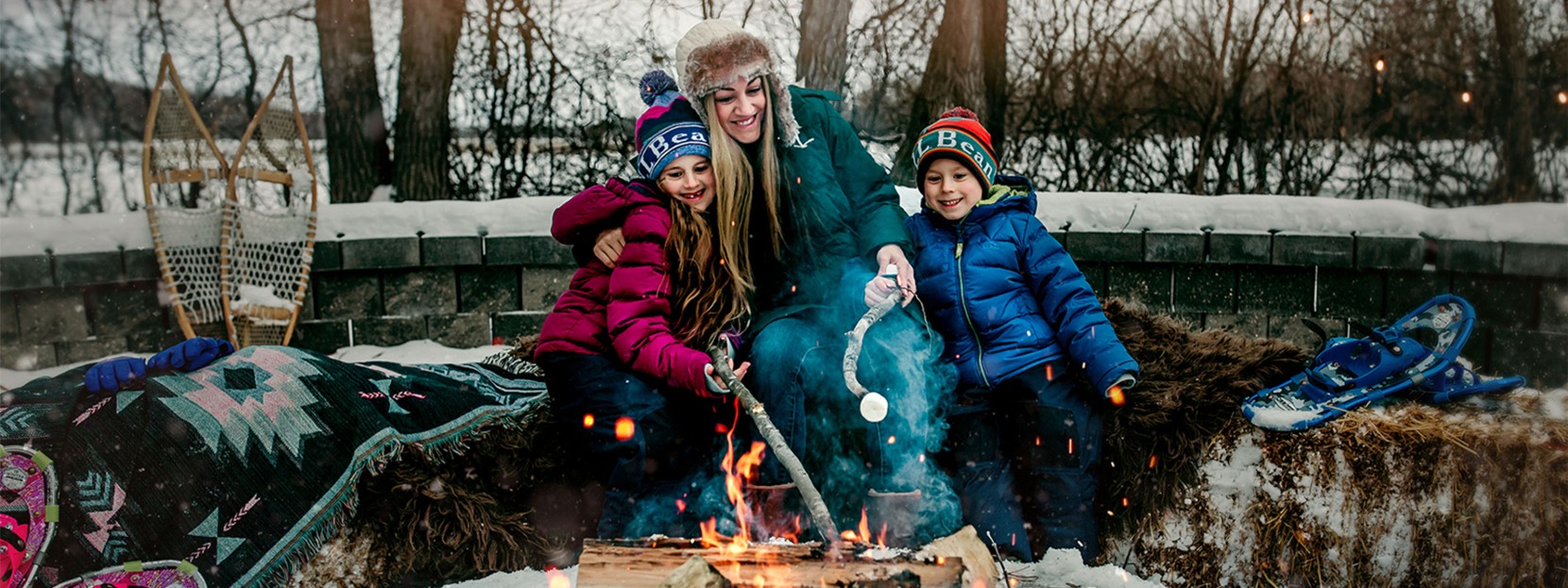 An outdoor fire on a snowy afternoon, a mom and 2 kids sitting on hay bales with blankets roasting marshmallows.