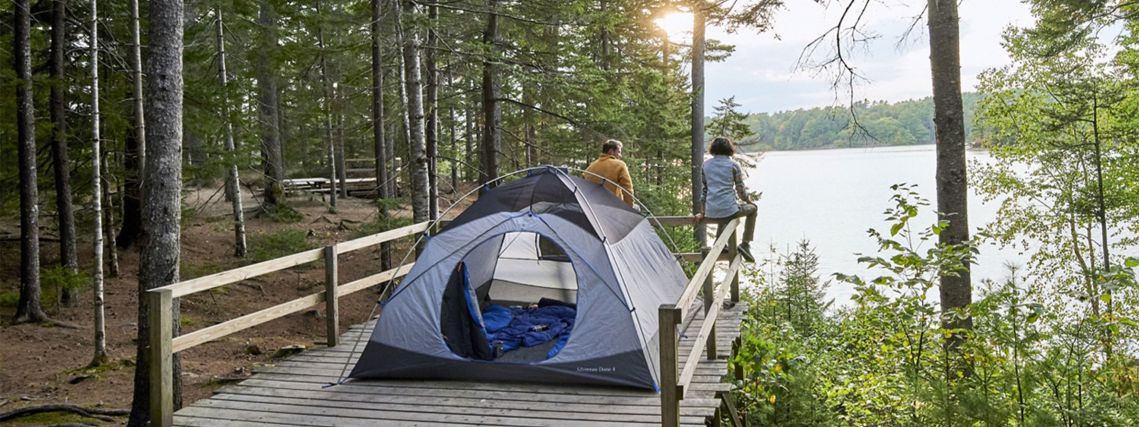 A couple sitting on a fence looking out at the water, their tent set up on a platform behind them.