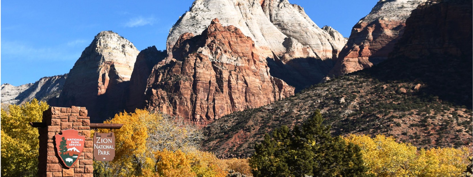A beautiful rock formation against a bright blue sky with a Zion National Park entrance sign in the foreground.