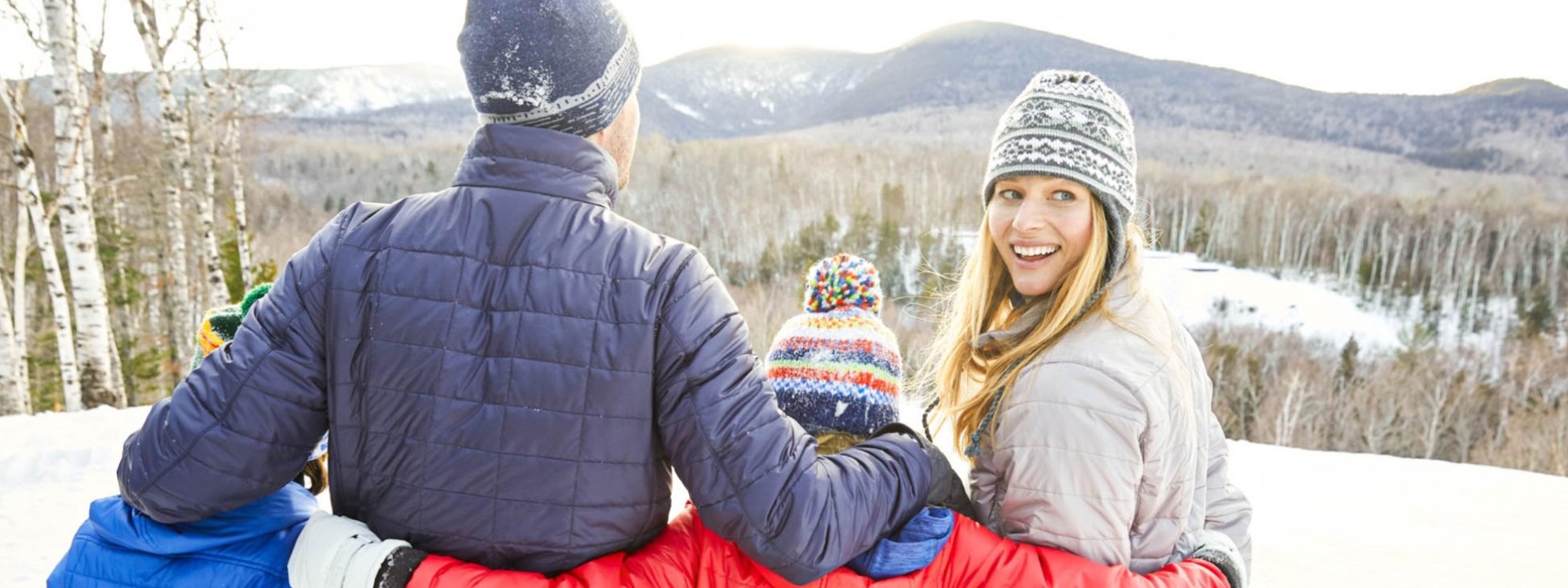 A family of 4 outside in winter, arms around each other, enjoying a beautiful mountain view. 