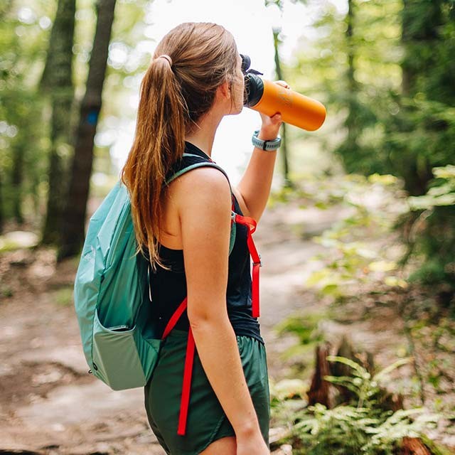 A young woman stops for a drink of water on a wooded trail.