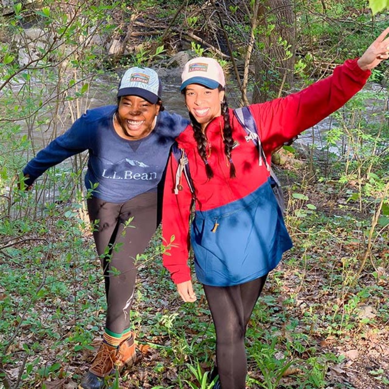2 smiling women walk toward the camera near a stream in the woods.