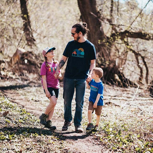 A dad holding hands with two young kids while walking on a wooded trail.