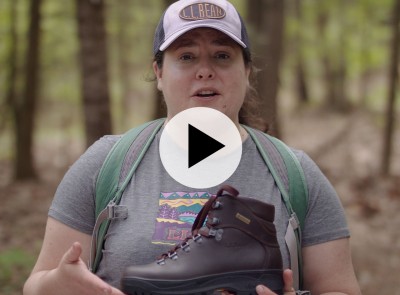 A woman wearing a backpack in the woods, looking at the camera and holding a hiking boot.