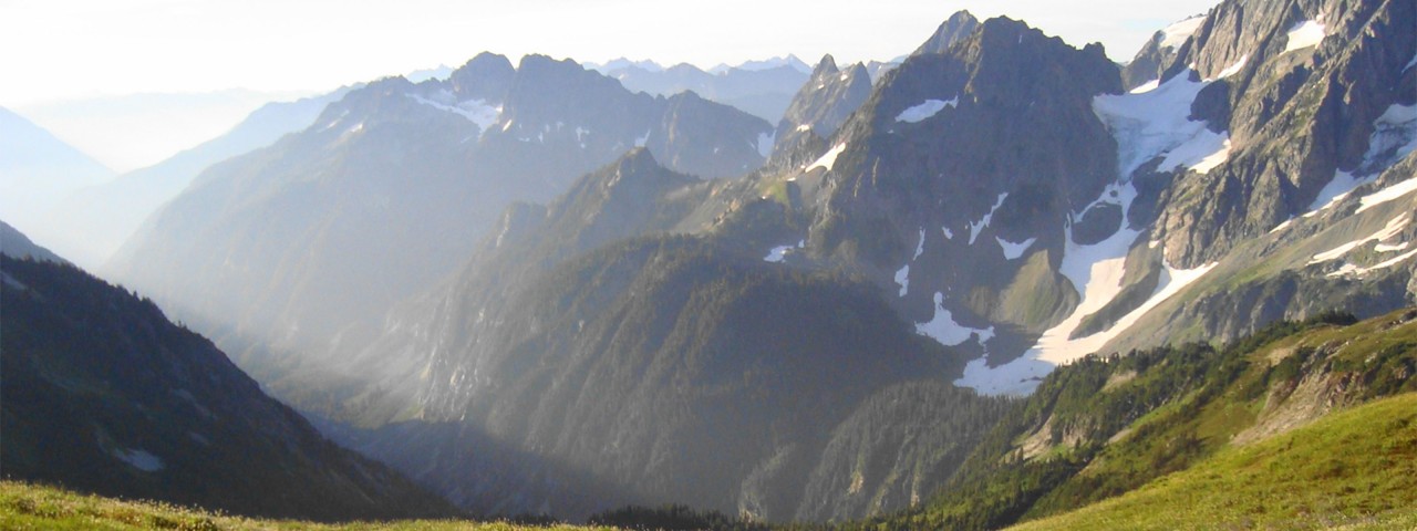 A green meadow in the foreground transitions to jagged, snow-covered mountains surrounding a steep valley.