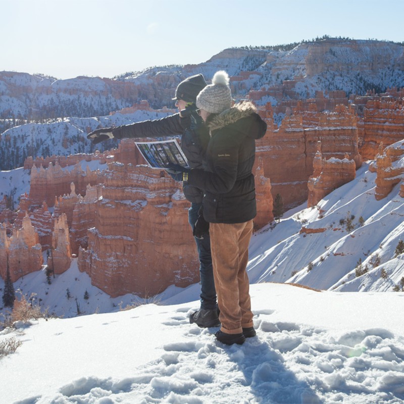 Two people in winter clothing stand at the edge of a snowy cliff pointing into distance where tall red rock spires and cliffs stand.