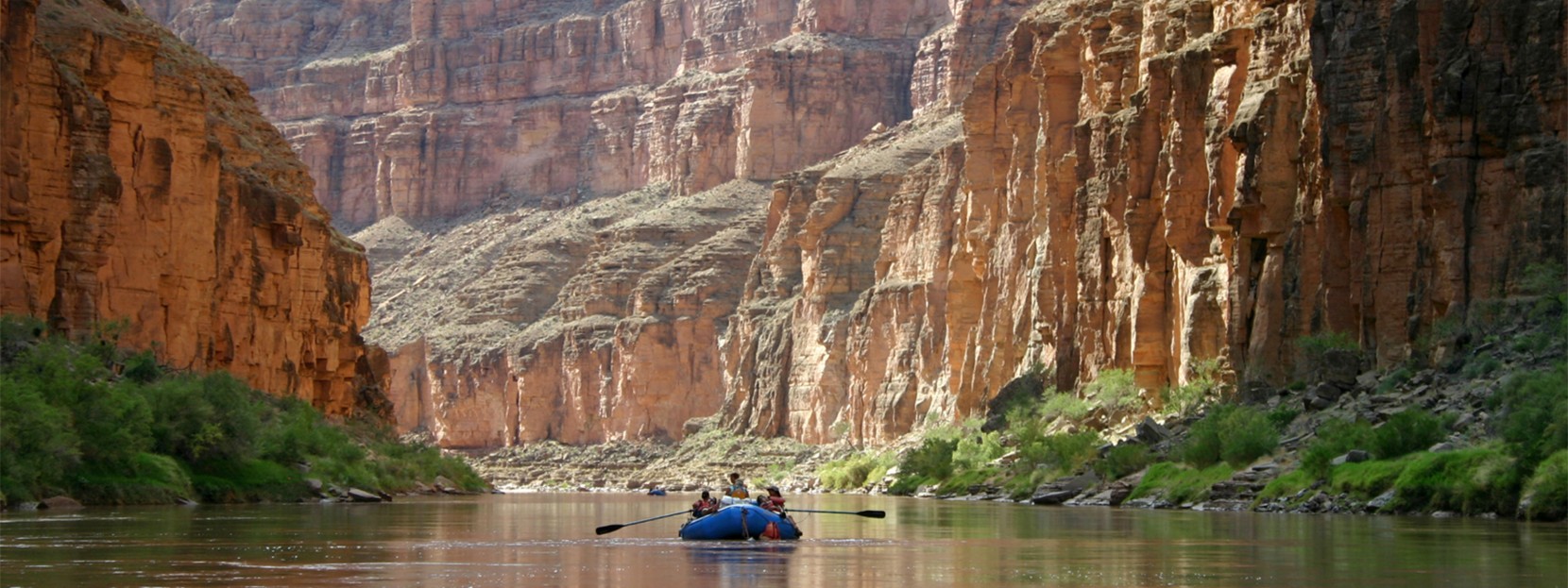 A raft with 3 people floats down a calm section of the Colorado River, dwarfed by towering canyon walls.
