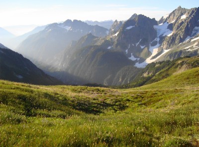 A green meadow in the foreground transitions to jagged, snow-covered mountains surrounding a steep valley.