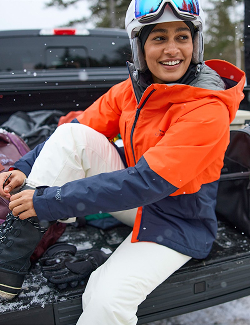 A woman wearing a ski helmet and goggles sits on a tailgate adjusting her boots.